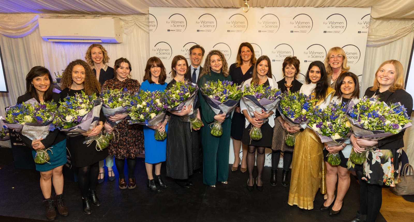 Shortlisted candidates, holding bunches of flowers, stand in a semi-circle with other guests.